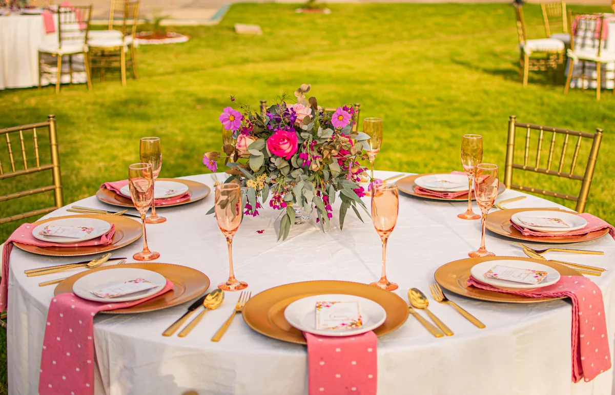 Mesa redonda con vajilla dorada, cristalería y centro floral en tonos rosa