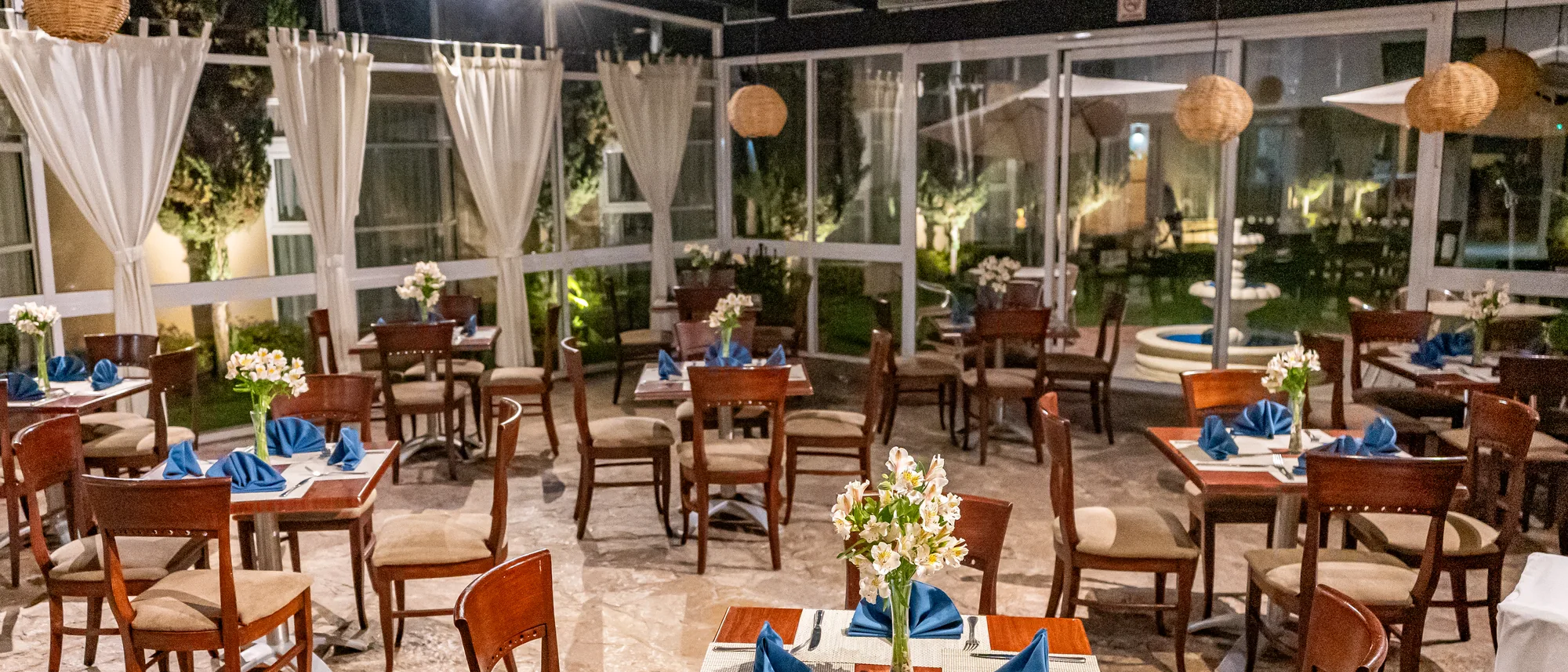Comedor del restaurante con muros de cristal, cortinas blancas y el jardín iluminado al fondo